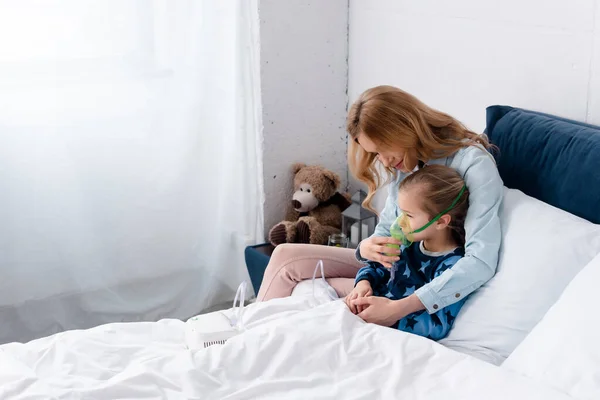 Madre tocando máscara respiratoria en hija asmática usando inhalador de compresor en el dormitorio - foto de stock