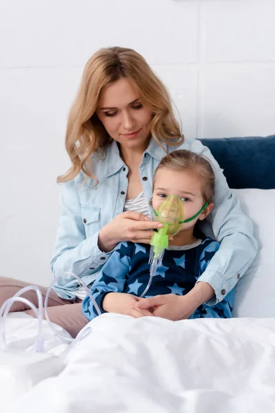 Schöne Mutter berührt Atemmaske auf kranke Tochter mit Kompressor-Inhalator im Schlafzimmer — Stockfoto