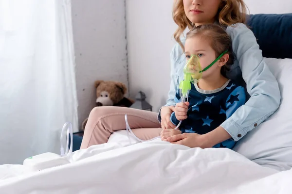 Madre sentada cerca de hija enferma en máscara respiratoria usando inhalador de compresor - foto de stock