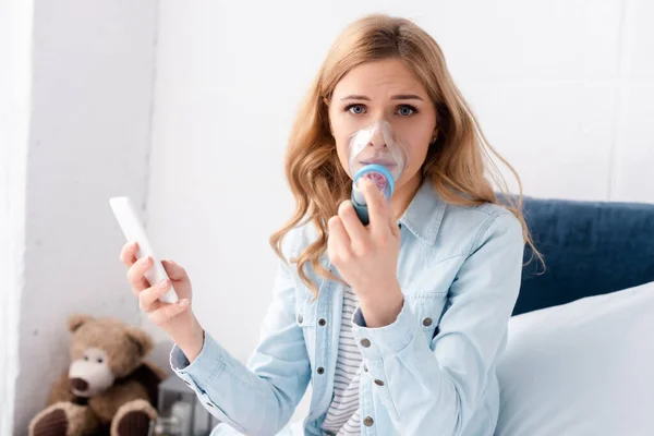 Selective focus of asthmatic woman using inhaler with spacer and holding smartphone — Stock Photo