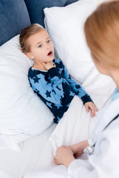 Selective focus of sick kid with opened mouth touching neck near doctor — Stock Photo