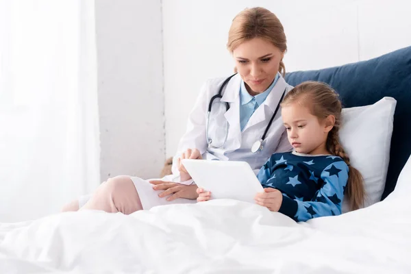 Atractivo médico de bata blanca sentado cerca de un niño enfermo con tableta digital - foto de stock
