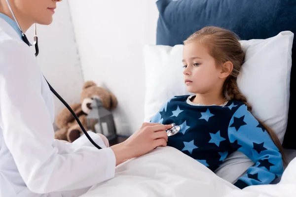 Doctor with stethoscope examining sick child in bedroom — Stock Photo