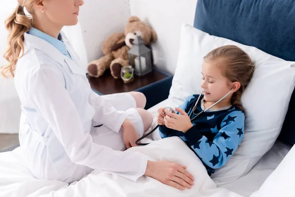 Доктор сидит на кровати рядом с больным ребенком со стетоскопом — стоковое фото