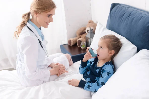 Happy doctor looking at asthmatic child with opened mouth using inhaler — Stock Photo