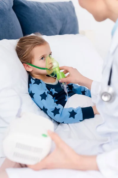 Médico de bata blanca tocando mascarilla respiratoria en niño asmático usando inhalador de compresor - foto de stock