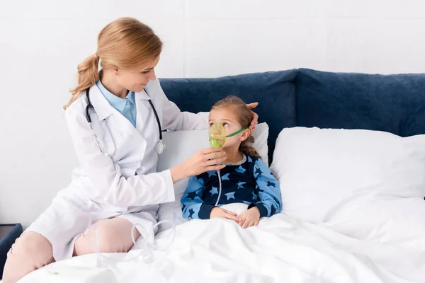Hermoso médico de bata blanca tocando máscara respiratoria en niño asmático - foto de stock