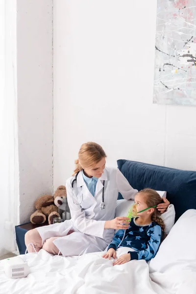 Médecin attrayant en manteau blanc touchant masque respiratoire sur enfant asthmatique — Photo de stock