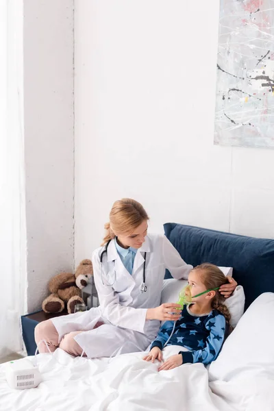 Attractive doctor in white coat touching respiratory mask on asthmatic child — Stock Photo