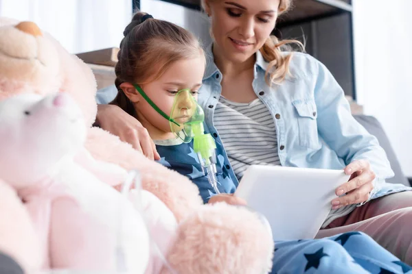 Selective focus of asthmatic kid using respiratory mask and holding digital tablet near mother and soft toys — Stock Photo