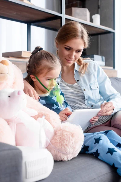 Selective focus of asthmatic kid using compressor inhaler and holding digital tablet near mother and soft toys — Stock Photo