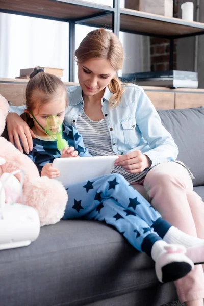 Selective focus of asthmatic child using compressor inhaler and holding digital tablet near mother and soft toy — Stock Photo