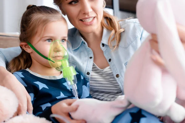 Selective focus of asthmatic kid using respiratory mask and looking at soft toy near mother — Stock Photo