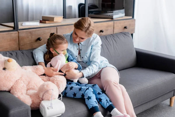 Niño asmático usando inhalador de compresor cerca de madre cariñosa con juguete suave - foto de stock