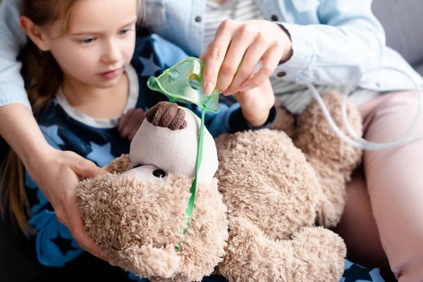 Selective focus of mother touching respiratory mask on teddy bear near sick kid — Stock Photo