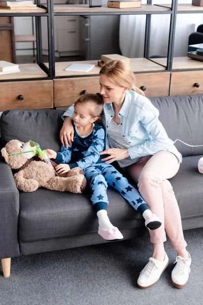 Kid touching respiratory mask on teddy bear near mother sitting on sofa — Stock Photo