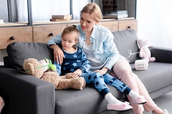 Kind schaut Atemmaske auf Teddybär neben Mutter — Stockfoto