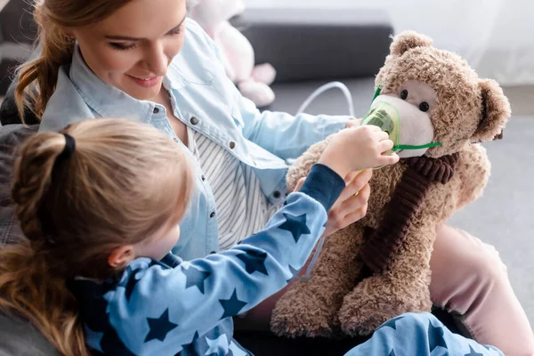 Selective focus of kid touching respiratory mask on teddy bear near happy mother — Stock Photo