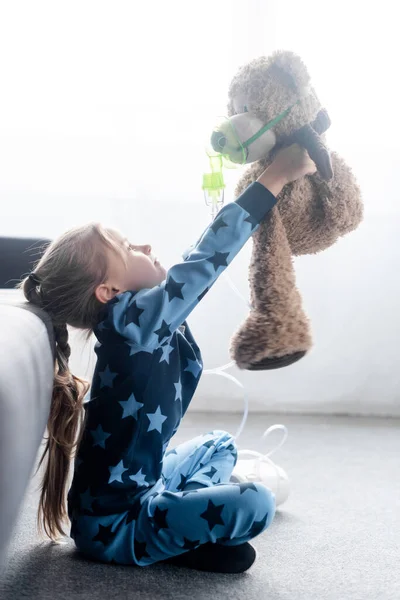 Vue latérale de mignon enfant tenant ours en peluche dans le masque respiratoire — Photo de stock