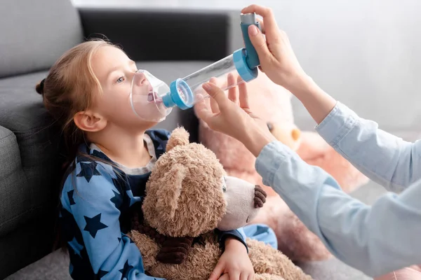 Madre che tiene l'inalatore con distanziatore vicino al bambino a casa — Foto stock