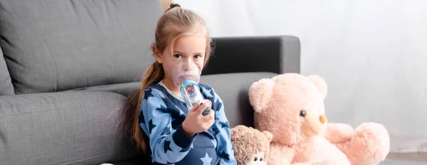 Panorámica de niño enfermo usando inhalador con espaciador cerca de juguetes blandos - foto de stock