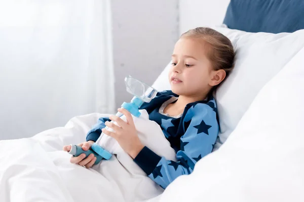 Больной ребенок держит ингалятор с прокладкой во время лежания в спальне — стоковое фото