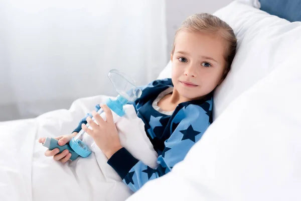 Niño asmático sosteniendo inhalador con espaciador mientras está acostado en el dormitorio - foto de stock