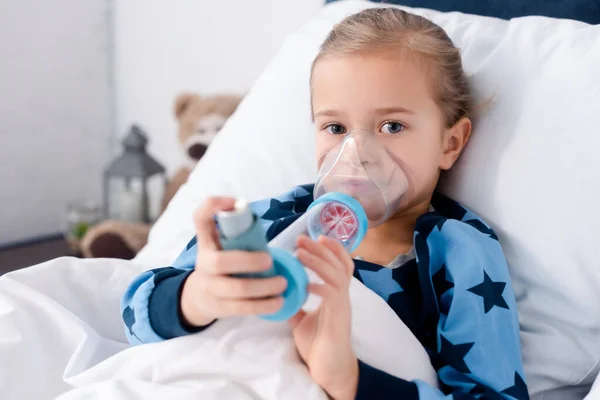 Enfoque selectivo del niño enfermo utilizando inhalador con espaciador y mirando a la cámara - foto de stock