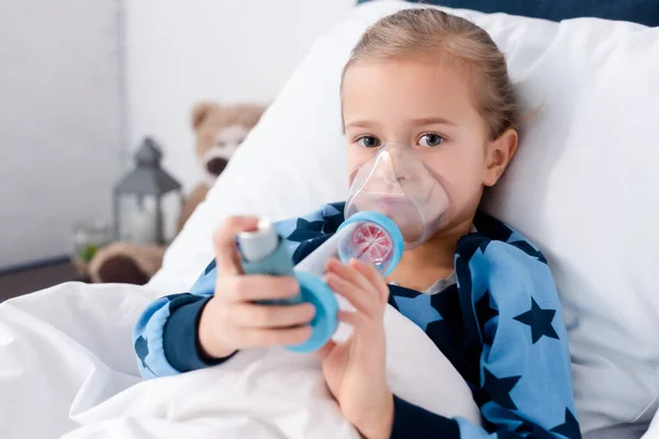 Selective focus of sick kid using inhaler with spacer and looking at camera — Stock Photo