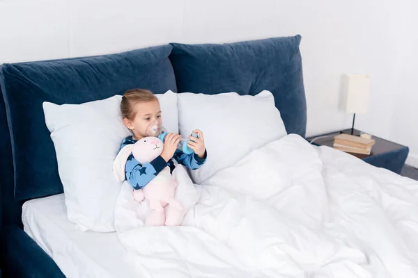 Niño enfermo usando inhalador con espaciador cerca de juguete de conejo - foto de stock