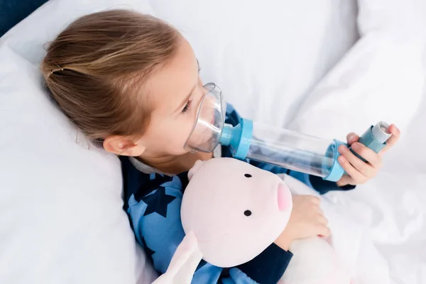 Niño enfermo usando inhalador con espaciador y sosteniendo juguete suave - foto de stock