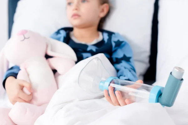 Selective focus of sick kid holding soft toy and inhaler with spacer — Stock Photo