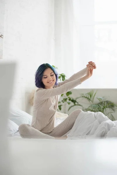 Selektiver Fokus einer Frau mit buntem Haar, ausgestreckten Händen und überkreuzten Beinen, die auf dem Bett lächelt — Stockfoto