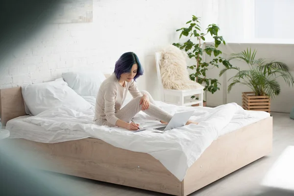 Enfoque selectivo del freelancer con la escritura colorida del pelo en el cuaderno cerca del ordenador portátil en la cama en el dormitorio - foto de stock