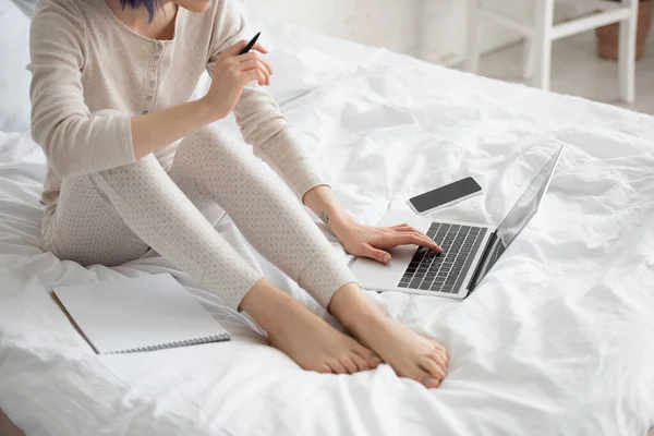 Vista recortada de freelancer con colorido cabello que sostiene la pluma y el trabajo en el ordenador portátil cerca de teléfono inteligente y copybook en la cama - foto de stock