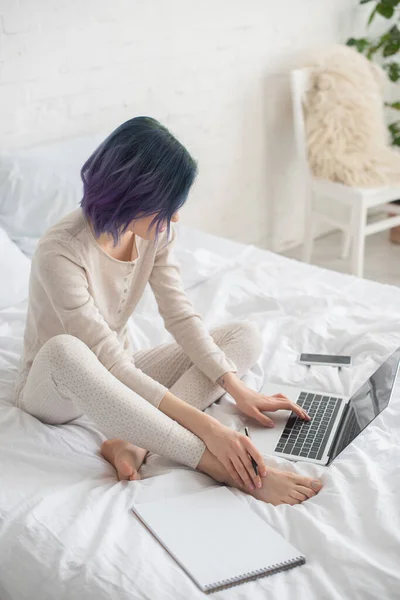 Visão de alto ângulo do freelancer com caneta de retenção de cabelo colorido e trabalhando em laptop perto de Copybook na cama — Fotografia de Stock