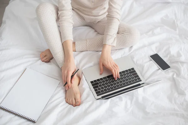 Partial view of freelancer with pen working on laptop near smartphone and notebook on bed — Stock Photo