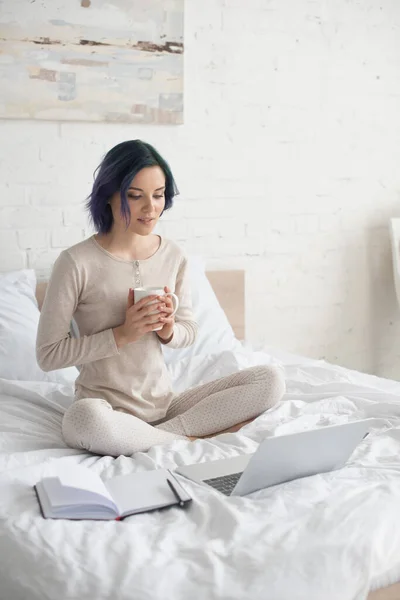 Libero professionista con capelli colorati e gambe incrociate in possesso di una tazza di tè e guardando computer portatile vicino notebook sul letto — Foto stock