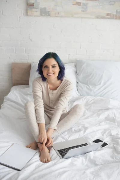 Высокоугольный вид фрилансера с цветными волосами, улыбающегося и смотрящего на камеру рядом с ноутбуком, ноутбуком и смартфоном на кровати — стоковое фото
