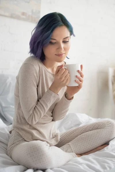Nachdenkliche Frau mit buntem Haar und überkreuzten Beinen, die eine Tasse Tee auf dem Bett im Schlafzimmer hält — Stockfoto