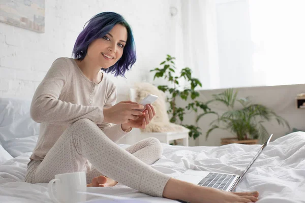 Freelancer com cabelo colorido sorrindo, olhando para a câmera e usando smartphone perto de laptop e xícara de chá na cama — Fotografia de Stock