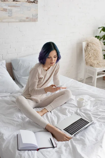 Vue grand angle de belle pigiste avec des cheveux colorés en utilisant un smartphone près d'un ordinateur portable et un ordinateur portable sur le lit dans la chambre — Photo de stock