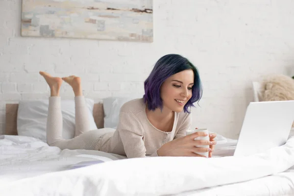 Freelancer com cabelo colorido e xícara de chá usando laptop, sorrindo e deitado na cama no quarto — Fotografia de Stock