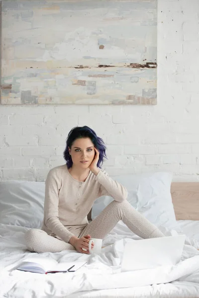 Freelancer with colorful hair and cup of tea looking at camera and sitting near laptop and copybook on bed — Stock Photo