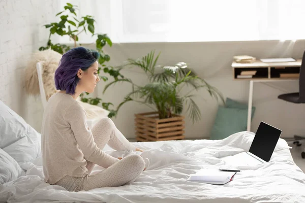 Freelancer con pelo colorido sentado cerca de la computadora portátil y portátil en la cama en el dormitorio - foto de stock