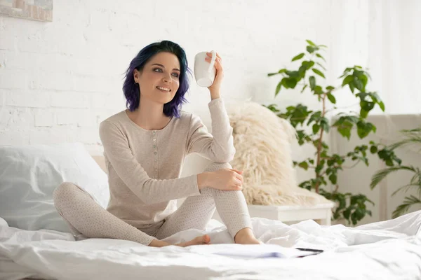 Mädchen mit buntem Haar hält Tasse Tee, lächelt und sitzt auf dem Bett im Schlafzimmer — Stockfoto