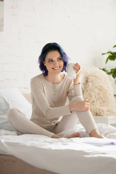 Mädchen mit buntem Haar und Tasse Tee lächelt und sitzt auf dem Bett im Schlafzimmer — Stockfoto