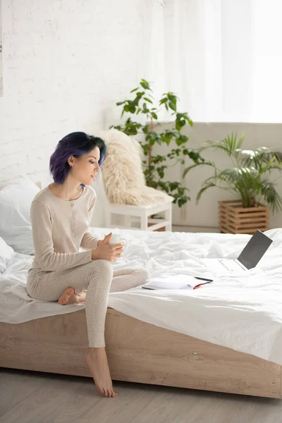 Freelancer com cabelo colorido e xícara de chá olhando para notebook perto de laptop na cama no quarto — Fotografia de Stock