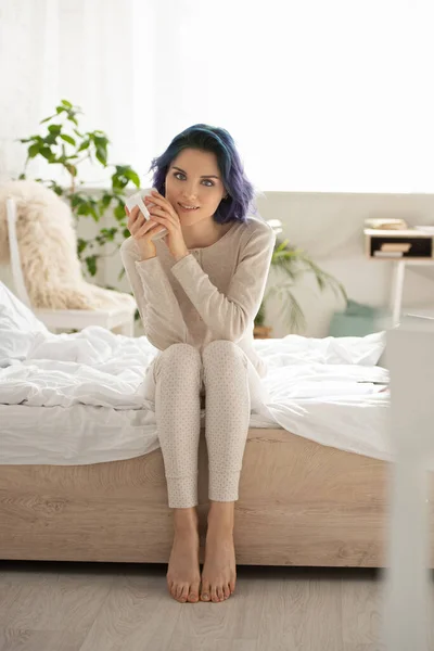 Menina com cabelo colorido e xícara de chá olhando para a câmera, sorrindo e sentado na cama no quarto — Fotografia de Stock