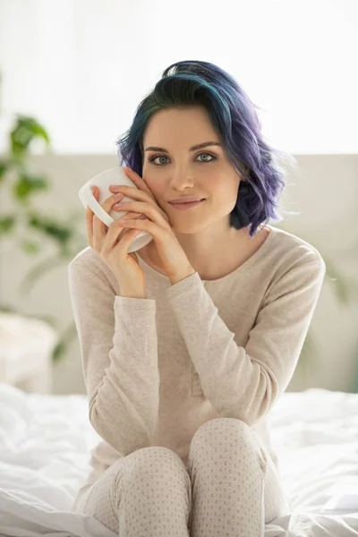 Девушка с раскрашенными волосами держит чашку чая, смотрит в камеру и улыбается на кровати в спальне — стоковое фото